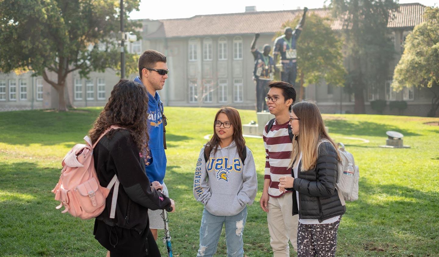 五名学生在圣何塞州立大学的高塔草坪上交谈，远处是胜利礼炮纪念碑.