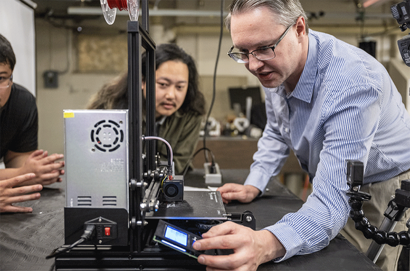 教授的照片. 汤姆·马杜拉向学生展示3D打印机的功能.